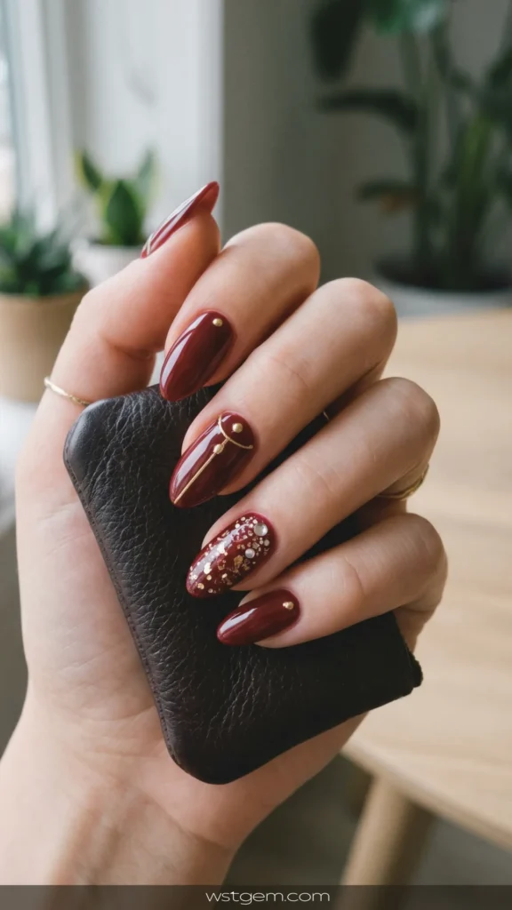 Close-up of a person's hand holding a brown leather wallet, showcasing a glossy dark red and gold manicure. The oval nails feature a deep maroon polish; the index, middle, and pinky fingers are adorned with delicate gold studs and lines, while the ring finger serves as an accent nail covered in gold and silver glitter flakes.